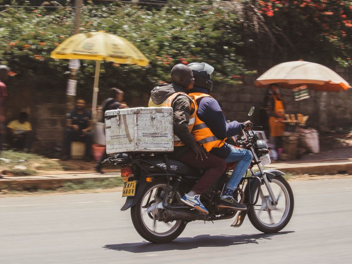 A delivery rider and passenger on a motorcycle, carrying a delivery box, moving through traffic